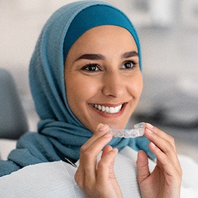 Woman in blue head wrap smiling in dental chair holding Invisalign